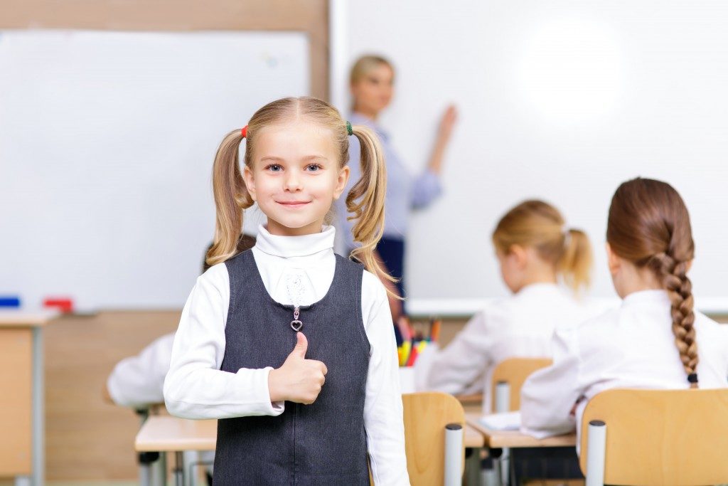 Little girl is smiling lovely and showing thumbs up while standing in the classroom
